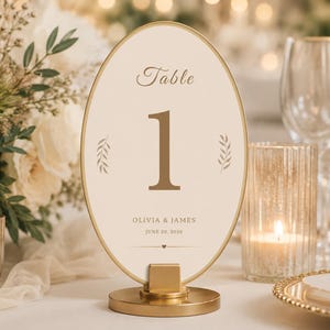 Op de afbeelding: Een ovaal tafelnummerbordje met een gouden frame en voet. De crèmekleurige kaart bevat de tekst "Table 1", de namen "Olivia & James" en de datum "June 20, 2028" in bruin lettertype, geaccentueerd door twee kleine bruine bladillustraties.