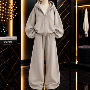 Puede incluir: Un conjunto de dos piezas en color beige que consta de una sudadera con capucha y cremallera de manga larga y pantalones anchos a juego. La sudadera tiene cierre de cremallera frontal y capucha, mientras que el pantalón tiene cordón.
