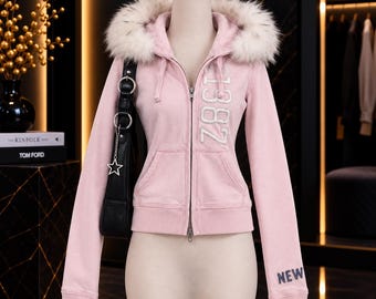 May include: A light pink zip-up hoodie featuring a white faux fur-trimmed hood, long sleeves, and two front pockets. The front displays the vertical text 1982 in white, while the left sleeve cuff features the word NEW in dark blue embroidery.