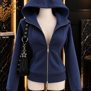 May include: A navy blue zip-up hoodie featuring a large hood, long sleeves with ribbed cuffs, and a ribbed hem. The garment has a central silver-toned metal zipper closure and vertical seam detailing along the front torso area.