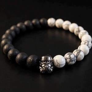 May include: A beaded bracelet featuring a split design of matte black porous beads and white marbled beads. A metallic silver-toned helmet charm, resembling a stormtrooper, is positioned at the centre, connecting the two distinct sections of beads.