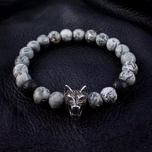 May include: A beaded bracelet featuring round, grey and white marbled stone beads interspersed with two textured black porous beads. A central silver-toned metal charm shaped like a detailed wolf head is positioned at the front of the bracelet.