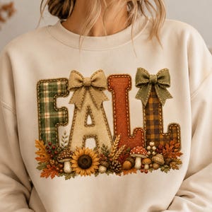Op de afbeelding: Een crèmekleurig sweatshirt met ronde hals en het woord FALL in grote, gestructureerde letters. De letters zijn versierd met groen ruitpatroon, beige jute, effen oranje en bruin ruitpatroon, geaccentueerd door twee beige en olijfgroene strikken.