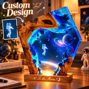 May include: A pentagonal resin and wood lamp featuring a space scene with an astronaut, planets, and a galaxy. The wooden base is engraved with the text "To the Moon" and a crescent moon icon. The resin glows with blue light.