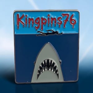 Può includere: Una spilla smaltata quadrata con una sezione superiore azzurra con il testo rosso Kingpins76 e una sagoma nera di un nuotatore. La sezione inferiore è blu scuro e mostra una testa di squalo grigia con la bocca aperta e denti bianchi.