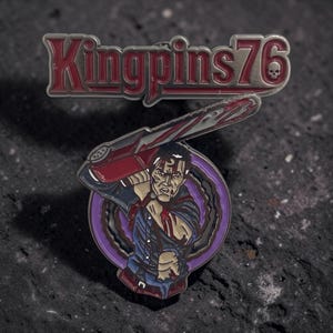 May include: A metal enamel pin featuring the text Kingpins76 in red and silver with a small skull icon. Below, a circular design depicts a character holding a red chainsaw over their shoulder, with blood splatters on their blue shirt and face.