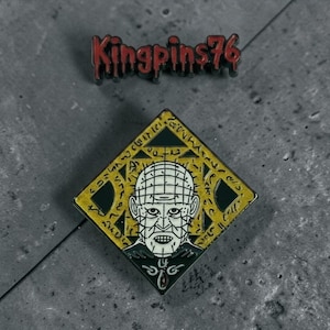 Puede incluir: Un pin de esmalte en forma de diamante con un retrato estilizado de una cabeza pálida con patrón de cuadrícula, centrado sobre un fondo geométrico dorado y negro con símbolos grabados. Sobre el pin, el texto Kingpins76 está en rojo.