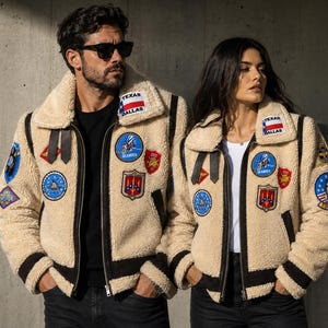 Könnte beinhalten: Zwei beigefarbene Bomberjacken aus Sherpa-Fleece mit dunkelbraunen Lederbesätzen, einem breiten Kragen und mehreren gestickten Aufnähern, darunter eine Texas-Dallas-Flagge, ein Seabees-Emblem und ein Tomcat-Abzeichen auf Brust und Ärmeln.