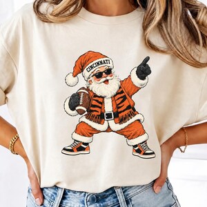 Peut inclure: Un t-shirt crème avec une illustration du Père Noël portant des lunettes de soleil, un bonnet avec le texte CINCINNATI et une veste à rayures de tigre. Le Père Noël tient un ballon de football, pointe vers le haut et porte des baskets.