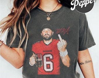 Puede incluir: Una camiseta de manga corta gris carbón con el gráfico de un jugador de fútbol americano con camiseta roja y el número 6, sosteniendo una jarra de cerveza y haciendo un gesto con el dedo medio. Incluye una firma roja y el texto Buccaneers.