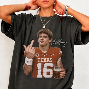 May include: A charcoal grey short-sleeve t-shirt featuring a graphic print of a football player in a burnt orange Texas jersey with the number 16, holding a glass of beer and giving the middle finger, with the text Arch Manning 16 printed nearby.
