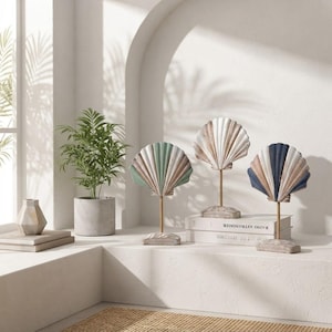 Peut inclure: Trois sculptures décoratives en forme de coquillage sur pied, chacune présentant une palette de couleurs unique en vert sauge, blanc et bleu marine, posées sur une surface blanche. Les coquillages sont montés sur des tiges métalliques.
