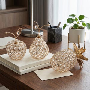 Pode incluir: Três esculturas decorativas de frutas feitas de fio de metal dourado e cristais facetados transparentes. O conjunto inclui uma maçã redonda, uma figura em forma de pera e um abacaxi, cada um com padrões de grade e hastes metálicas.