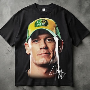 Puede incluir: Una camiseta negra de manga corta con un gran estampado gráfico de una persona que lleva una gorra de béisbol amarilla y verde con el texto U CAN'T SEE ME. Una firma blanca estilizada está impresa debajo del retrato en la parte inferior derecha.