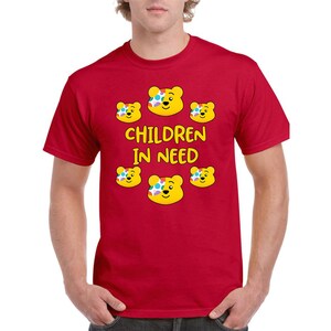 May include: A bright red short-sleeved t-shirt featuring a graphic print of five yellow bear faces with colourful polka-dot patches over one eye. The text "CHILDREN IN NEED" is printed in yellow capital letters in the centre of the bear faces.