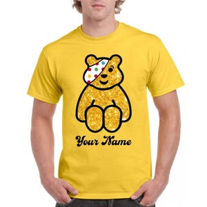 May include: A bright yellow short-sleeved t-shirt featuring a central graphic of a gold glitter teddy bear with a white eye patch covered in colourful polka dots. Below the bear, the black text reads Your Name in a cursive script font.