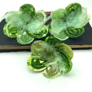 May include: Three green sequin flower embellishments with white feathers. The flowers are made of green sequins and have a three-dimensional shape.