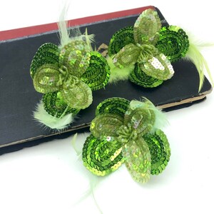 May include: Three green sequin flower embellishments with feather accents. The flowers are made of overlapping sequins in a light green color.