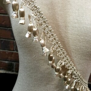 Peut inclure: Une garniture beige avec un motif de dentelle d&eacute;licat et une frange de petits glands. La garniture est pr&eacute;sent&eacute;e sur un mannequin recouvert de tissu.