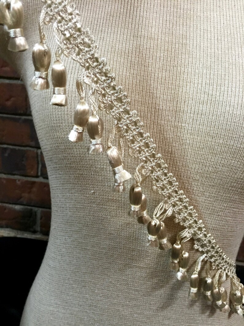 Peut inclure: Une garniture beige avec un motif de dentelle d&eacute;licat et une frange de petits glands. La garniture est pr&eacute;sent&eacute;e sur un mannequin recouvert de tissu.