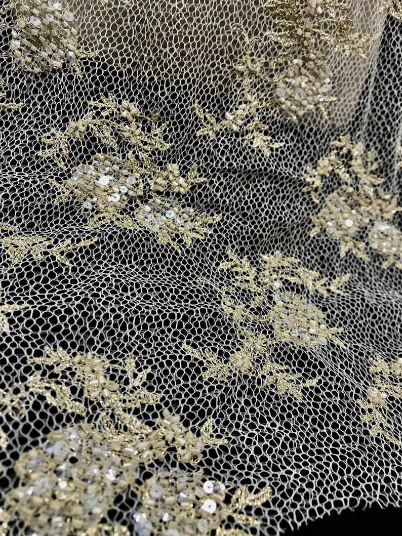 K&ouml;nnte beinhalten: Nahaufnahme eines elfenbeinfarbenen Spitzenstoffs mit aufwendiger floraler Stickerei. Das filigrane Design weist kleine, schimmernde Pailletten und Perlen auf, die ein strukturiertes und elegantes Aussehen erzeugen. Die Spitze ist vor einem dunklen Hintergrund platziert, der das detaillierte Muster hervorhebt.