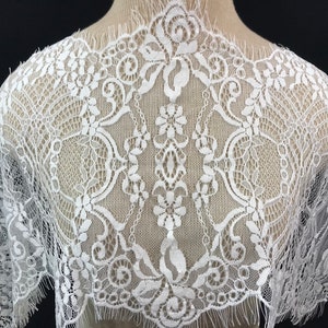May include: White lace fabric with a delicate floral pattern. The lace has a scalloped edge and a fringe.