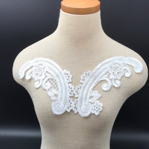 Op de afbeelding: Witte kantversiering met een bloemenpatroon. De versiering is V-vormig en perfect om een ​​toets van elegantie aan elk kledingstuk toe te voegen.