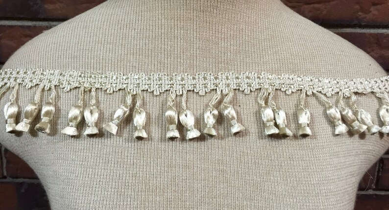 Peut inclure: Gros plan d'une garniture cr&egrave;me avec un motif de dentelle d&eacute;licat et une frange de petits glands.