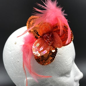Puede incluir: Un clip de pelo con forma de flor de color rosa coral y naranja con lentejuelas y plumas.