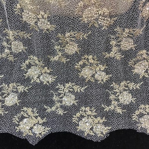 K&ouml;nnte beinhalten: Nahaufnahme eines zarten, cremefarbenen Spitzenstoffs mit floralem Muster. Die Spitze weist detaillierte Stickereien in hellem Gold auf, mit einem gewellten Rand. Das Design umfasst Blumengruppen und Bl&auml;tter, die eine elegante Textur erzeugen.