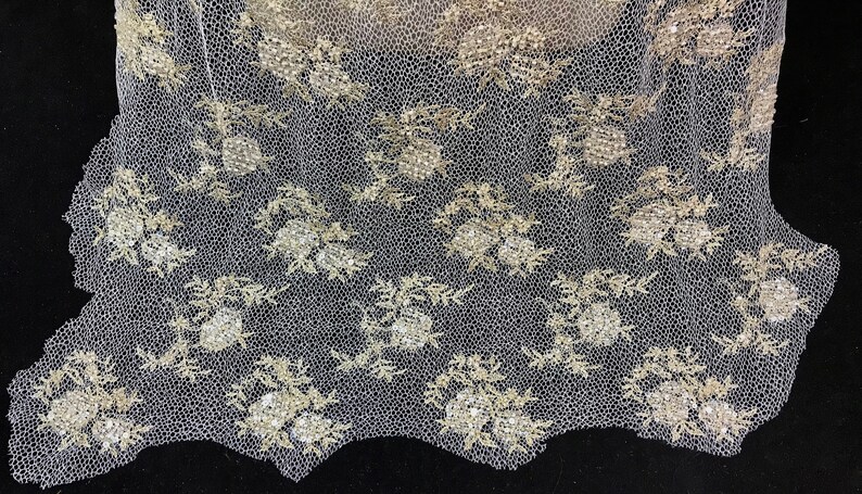 K&ouml;nnte beinhalten: Nahaufnahme eines zarten, cremefarbenen Spitzenstoffs mit floralem Muster. Die Spitze weist detaillierte Stickereien in hellem Gold auf, mit einem gewellten Rand. Das Design umfasst Blumengruppen und Bl&auml;tter, die eine elegante Textur erzeugen.