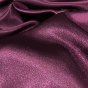 May include: Close-up of a deep purple satin fabric with a smooth, silky texture. The fabric is draped and folded, creating soft, flowing lines.
