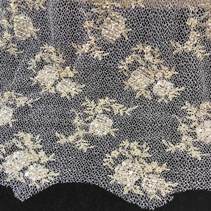 K&ouml;nnte beinhalten: Nahaufnahme eines wei&szlig;en Spitzenstoffs mit floralem Muster. Das Design zeigt gestickte Blumen mit Pailletten-Details in hellem Gold. Die Spitze hat ein zartes, offenes Gewebe und der Rand ist gewellt.