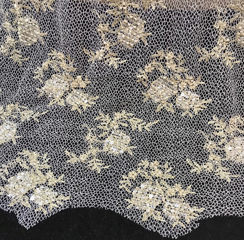 K&ouml;nnte beinhalten: Nahaufnahme eines wei&szlig;en Spitzenstoffs mit floralem Muster. Das Design zeigt gestickte Blumen mit Pailletten-Details in hellem Gold. Die Spitze hat ein zartes, offenes Gewebe und der Rand ist gewellt.
