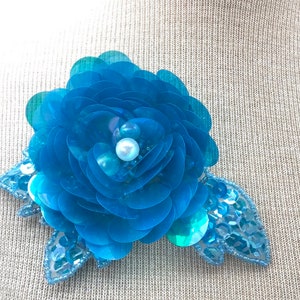 Può includere: Un fiore di tessuto blu turchese con un centro di perla e paillettes iridescenti. Il fiore è attaccato a una foglia di tessuto.