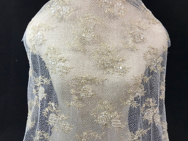 K&ouml;nnte beinhalten: Elfenbeinfarbener Spitzenstoff mit floralem Muster und schimmernden Pailletten. Der zarte Netzstoff ist &uuml;ber eine Schaufensterpuppe drapiert und zeigt das filigrane Design. Geeignet f&uuml;r Brautmode und Kleidung f&uuml;r besondere Anl&auml;sse.