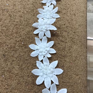 May include: A line of white, daisy-shaped floral embellishments. Each flower has multiple petals and a textured centre. The flowers are arranged vertically against a brown, textured surface, creating a decorative trim.