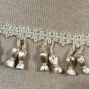 Peut inclure: Gros plan sur un tissu beige avec une garniture d&eacute;corative de dentelle blanche et de glands. La garniture pr&eacute;sente un motif r&eacute;p&eacute;titif de petits carr&eacute;s et un bord festonn&eacute;.