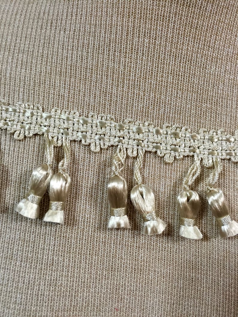 Peut inclure: Gros plan sur un tissu beige avec une garniture d&eacute;corative de dentelle blanche et de glands. La garniture pr&eacute;sente un motif r&eacute;p&eacute;titif de petits carr&eacute;s et un bord festonn&eacute;.