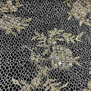 K&ouml;nnte beinhalten: Nahaufnahme eines elfenbeinfarbenen Spitzenstoffs mit goldfarbenen Blumenstickereien und Perlenakzenten. Die zarte netzartige Textur ist vor einem schwarzen Hintergrund sichtbar und unterstreicht das Design und die Handwerkskunst.
