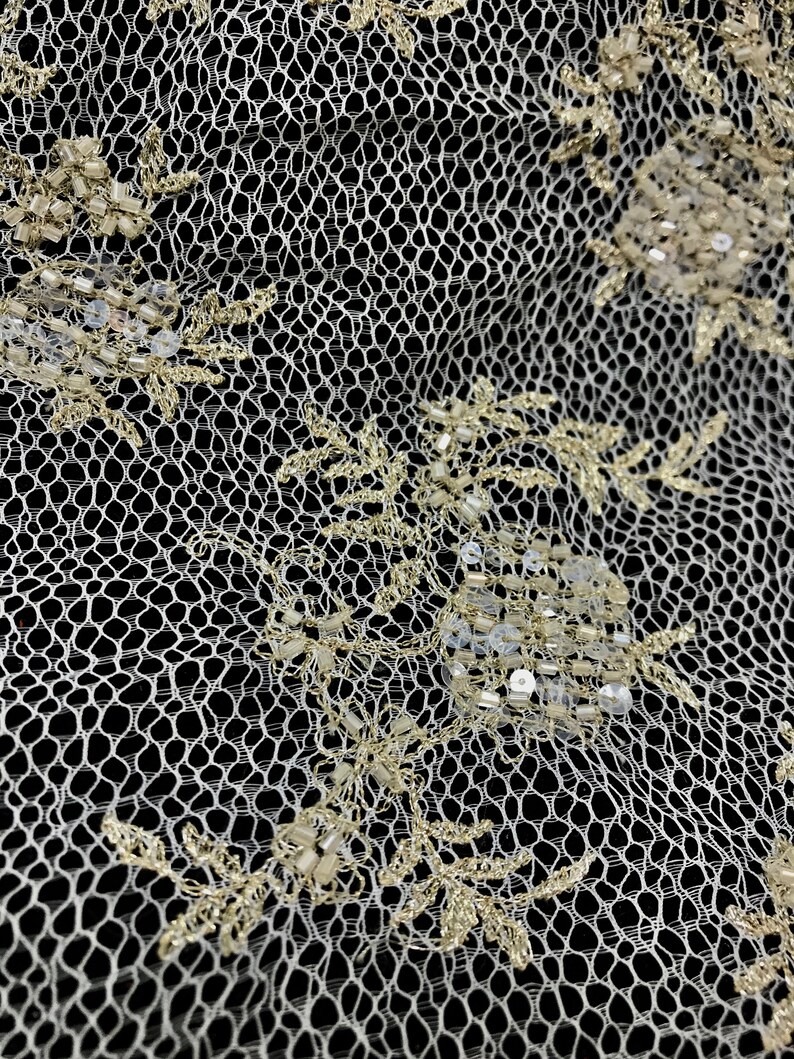 K&ouml;nnte beinhalten: Nahaufnahme eines elfenbeinfarbenen Spitzenstoffs mit goldfarbenen Blumenstickereien und Perlenakzenten. Die zarte netzartige Textur ist vor einem schwarzen Hintergrund sichtbar und unterstreicht das Design und die Handwerkskunst.