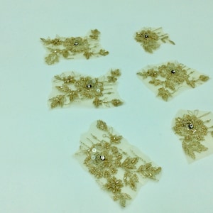 Puede incluir: Seis aplicaciones de encaje de cuentas doradas con centros de diamantes de imitación transparentes. Las aplicaciones están sobre un fondo blanco.