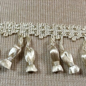 Peut inclure: Gros plan sur une garniture beige avec des glands. La garniture a un motif tiss&eacute; et les glands sont faits d'un mat&eacute;riau doux et soyeux.