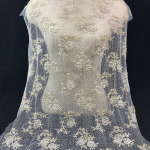 K&ouml;nnte beinhalten: Elfenbeinfarbener Spitzenstoff mit floralen und Pailletten-Verzierungen. Die zarte Spitze ist &uuml;ber eine Schaufensterpuppe drapiert und zeigt das aufwendige Design. Der Stoff weist ein sich wiederholendes Blumenmuster mit schimmernden Pailletten auf.