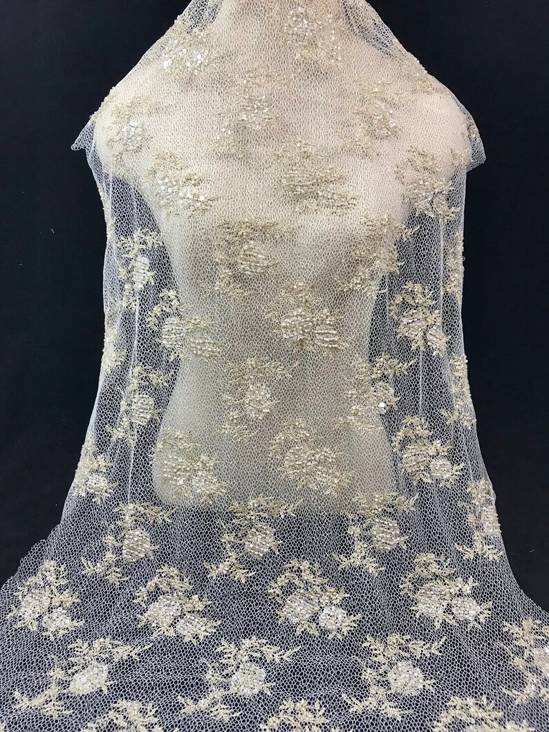 K&ouml;nnte beinhalten: Elfenbeinfarbener Spitzenstoff mit floralen und Pailletten-Verzierungen. Die zarte Spitze ist &uuml;ber eine Schaufensterpuppe drapiert und zeigt das aufwendige Design. Der Stoff weist ein sich wiederholendes Blumenmuster mit schimmernden Pailletten auf.