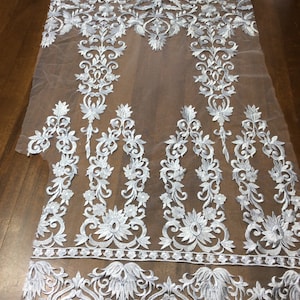 Peut inclure: Tissu de dentelle blanche brodée avec un motif floral sur un fond de tulle transparent. La dentelle a un design délicat et complexe avec un motif répétitif de fleurs et de feuilles.