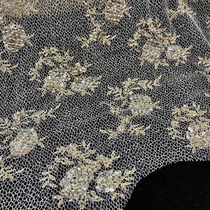 K&ouml;nnte beinhalten: Nahaufnahme eines elfenbeinfarbenen Spitzenstoffs mit Blumen- und Bl&auml;ttermustern. Die Spitze weist gestickte Blumen mit Pailletten-Details auf. Der Hintergrund ist schwarz und hebt das filigrane Design der Spitze hervor.