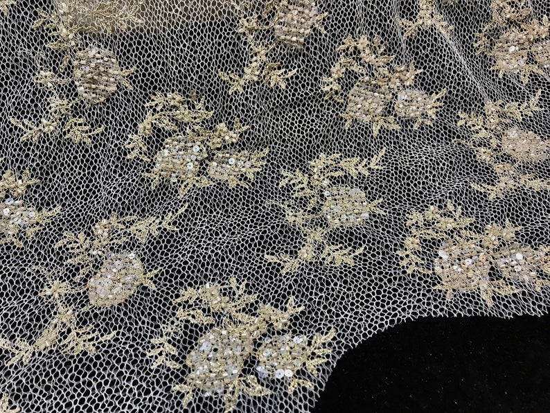 K&ouml;nnte beinhalten: Nahaufnahme eines elfenbeinfarbenen Spitzenstoffs mit Blumen- und Bl&auml;ttermustern. Die Spitze weist gestickte Blumen mit Pailletten-Details auf. Der Hintergrund ist schwarz und hebt das filigrane Design der Spitze hervor.