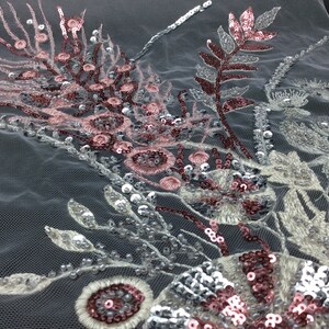 Può includere: Primo piano di un tessuto delicato ricamato con paillettes argentate e rosa. Il tessuto presenta un motivo floreale con foglie e fiori.