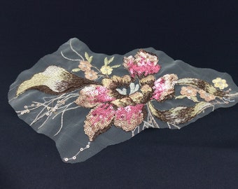May include: A delicate floral applique with pink, gold, and brown sequins on a sheer mesh fabric. The applique features a large flower with smaller flowers and leaves surrounding it.